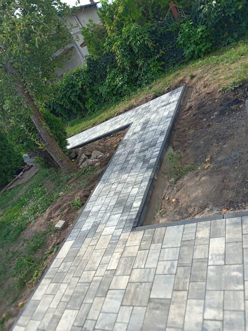 Ścieżka z szarej kostki brukowej ułożona na zboczu, widoczna z góry, z drzewem i zielenią w tle.