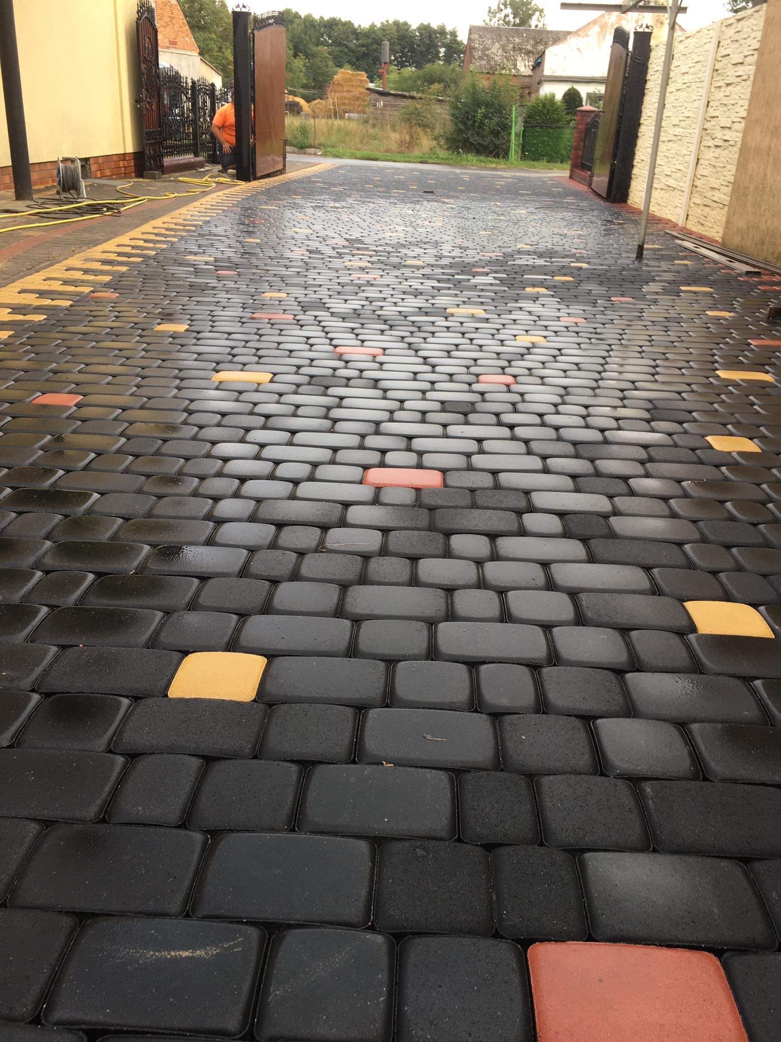 Ułożona kostka granitowa w kolorach szarym, żółtym i czerwonym, prowadząca do otwartej, metalowej bramy wjazdowej, widok z dołu.