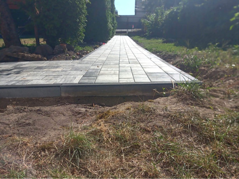 Ścieżka z kostki granitowej w ogrodzie, widok z poziomu gruntu, świeżo ułożona, z widoczną ziemią i trawą przy krawędziach.