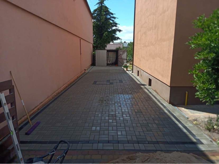 Ułożona kostka granitowa w wąskim przejściu między budynkami, widoczne narzędzia brukarskie: poziomica, szczotka i taczka oraz piasek na brzegu.