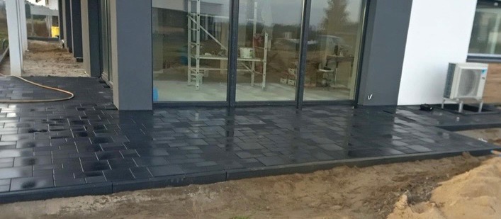 Świeżo ułożona, ciemnoszara kostka brukowa na tarasie przed nowoczesnym budynkiem z dużymi oknami przesuwnymi, widoczny jeszcze piasek obok krawężnika.