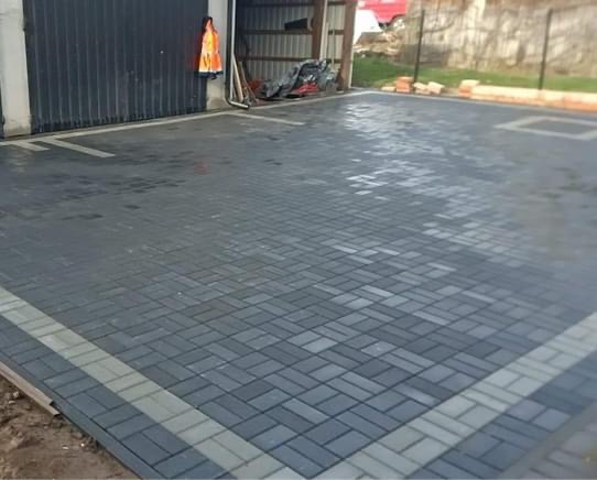 Ułożona kostka brukowa w odcieniach szarości na posesji przed garażem z pomarańczową kamizelką odblaskową wiszącą na ścianie.