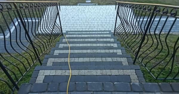 Schody zewnętrzne wyłożone kostką granitową w dwóch odcieniach szarości, z ozdobną, metalową balustradą w kolorze czarnym i złotym wykończeniem poręczy. Na schodach leży żółty kabel.