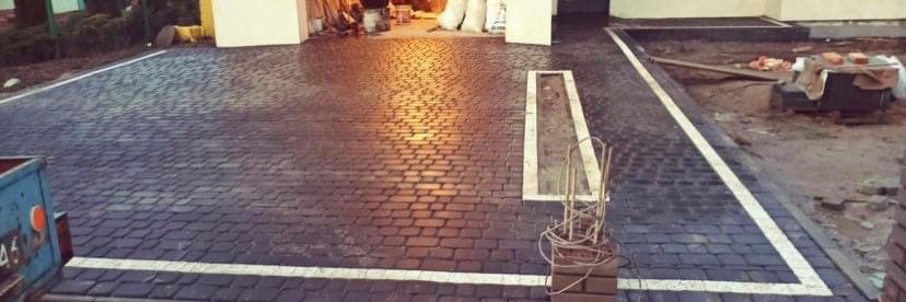 Ułożona kostka granitowa w kolorze grafitowym z białymi liniami wyznaczającymi miejsca parkingowe, fragmenty niedokończonej nawierzchni z widocznymi materiałami budowlanymi w tle.