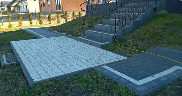 Ułożona kostka granitowa w odcieniach szarości tworząca chodnik i schody przed domem z czarną balustradą, widoczny trawnik i fragment elewacji budynku