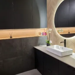 Nowoczesna łazienka z czarnymi szafkami, białą prostokątną umywalką i okrągłym lustrem z podświetleniem LED, wzorzysta tapeta w odcieniach beżu i brązu, oraz elementy dekoracyjne.