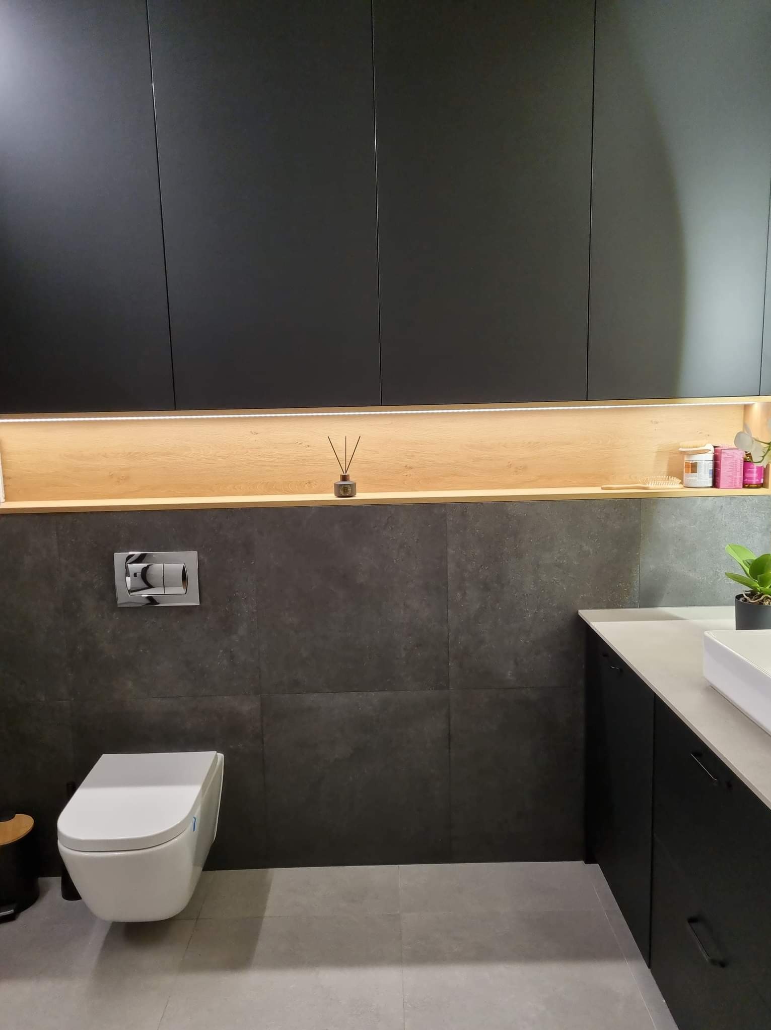 Nowoczesna łazienka z wiszącą toaletą, szarą płytką na ścianie, czarnymi szafkami i drewnianą wnęką z oświetleniem LED oraz dekoracjami.
