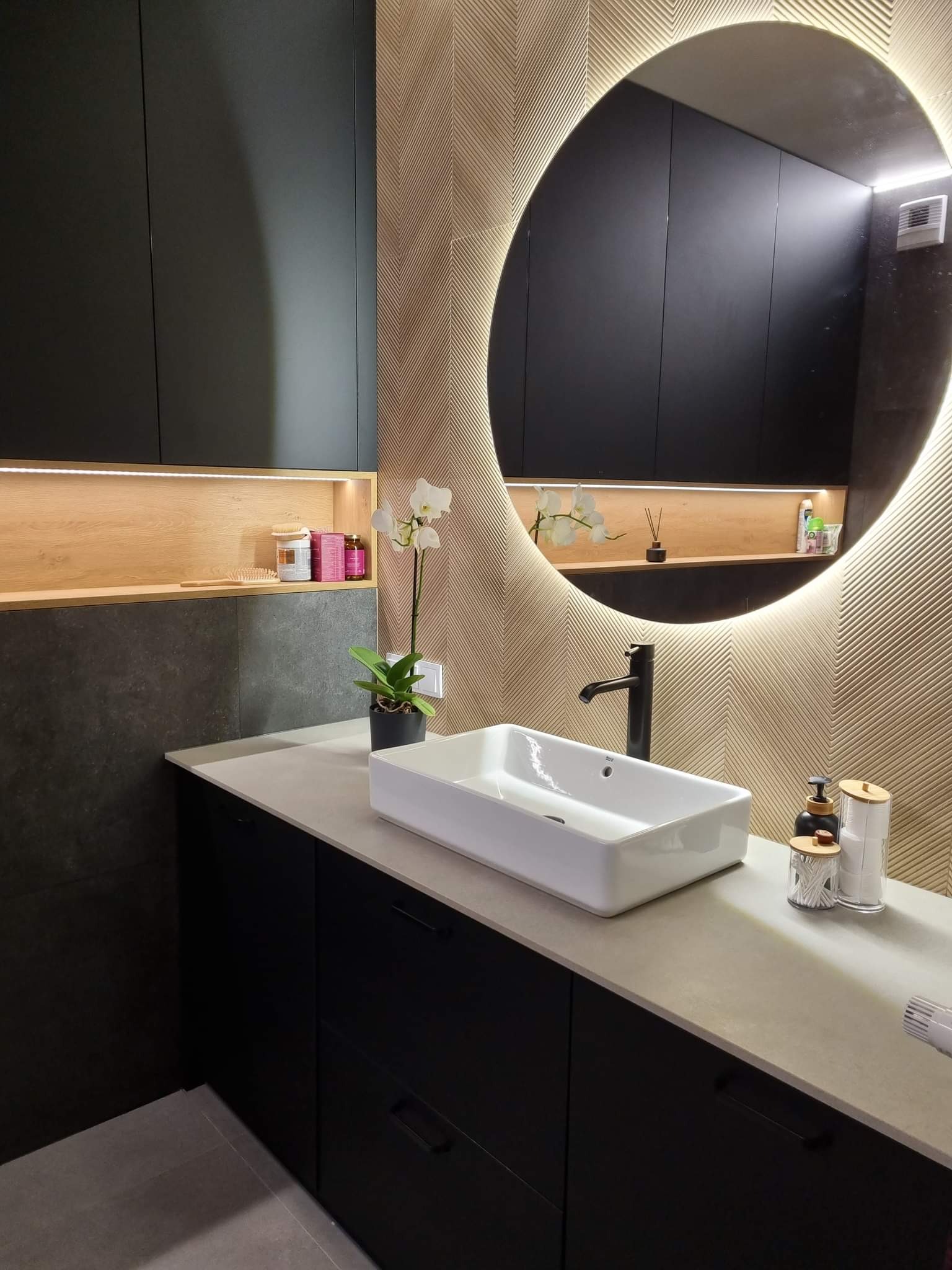 Nowoczesna łazienka z czarnymi szafkami, prostokątną białą umywalką nablatową, podświetlanym okrągłym lustrem i ścianą z dekoracyjną fakturą w jodełkę.
