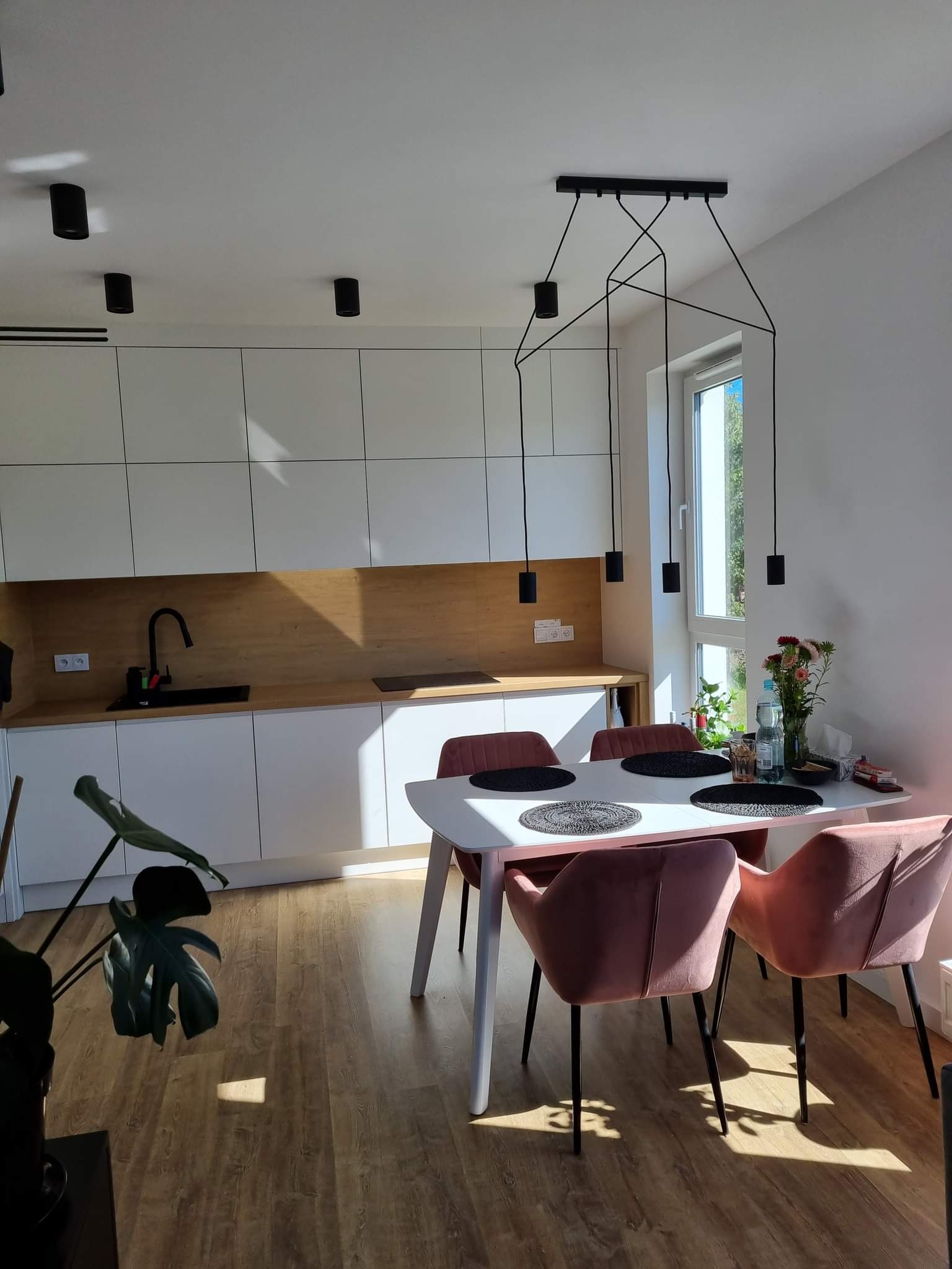 Jasna kuchnia z drewnianym blatem, białymi szafkami, czarną armaturą i stołem z różowymi krzesłami, oświetlona nowoczesną lampą wiszącą i punktowymi reflektorami.