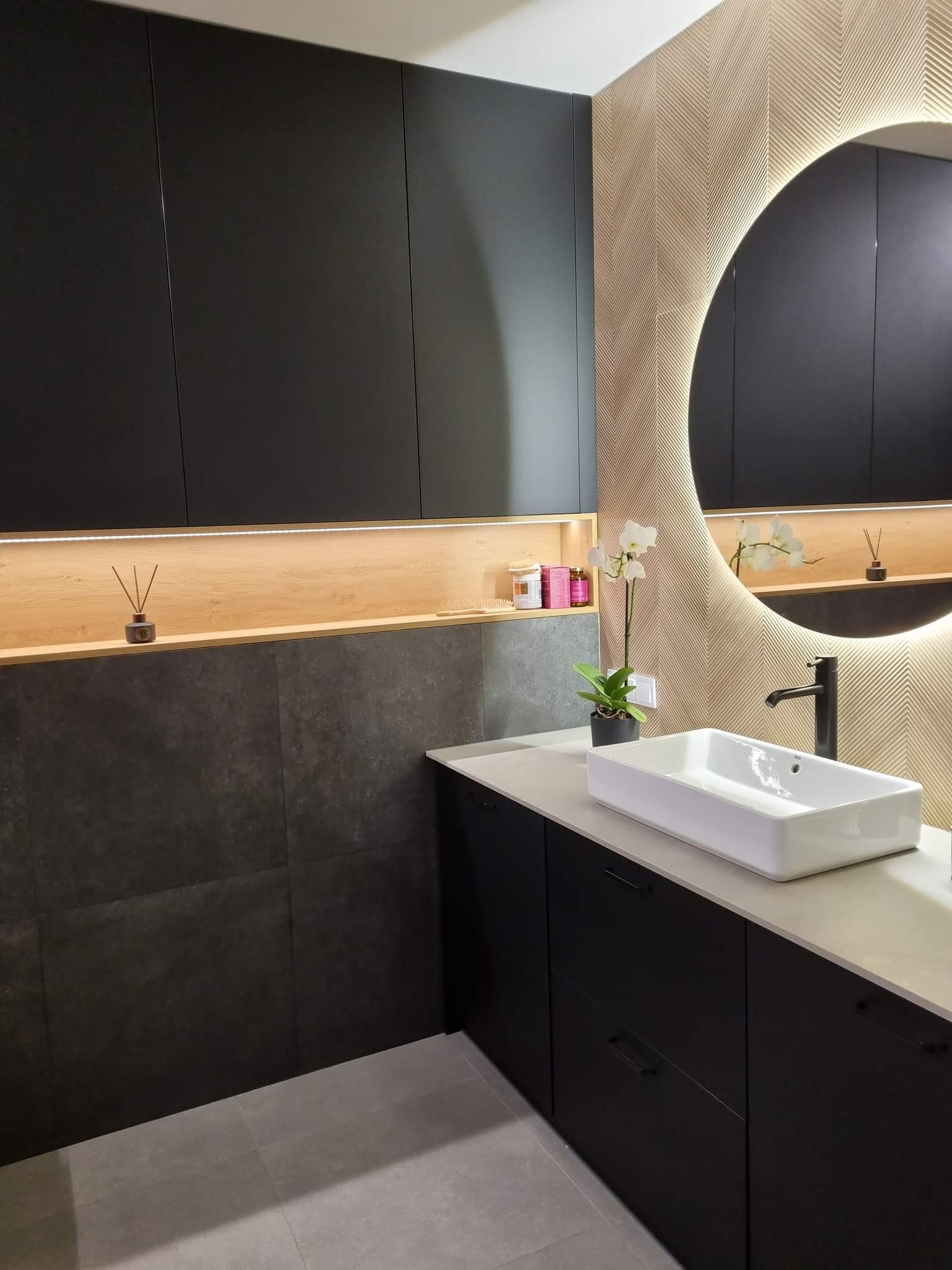 Nowoczesna łazienka z czarnymi szafkami, białą prostokątną umywalką i okrągłym lustrem z podświetleniem LED, wzorzysta tapeta w odcieniach beżu i brązu, oraz elementy dekoracyjne.