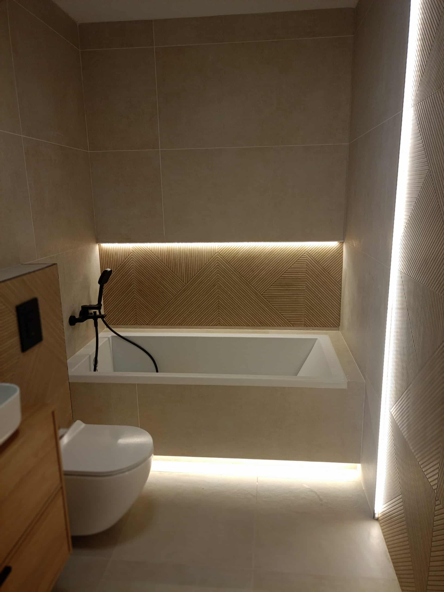 Nowoczesna łazienka z beżowymi płytkami, podświetlaną wanną i geometrycznym wzorem na ścianie. Minimalistyczny design z wiszącą toaletą i drewnianą szafką.