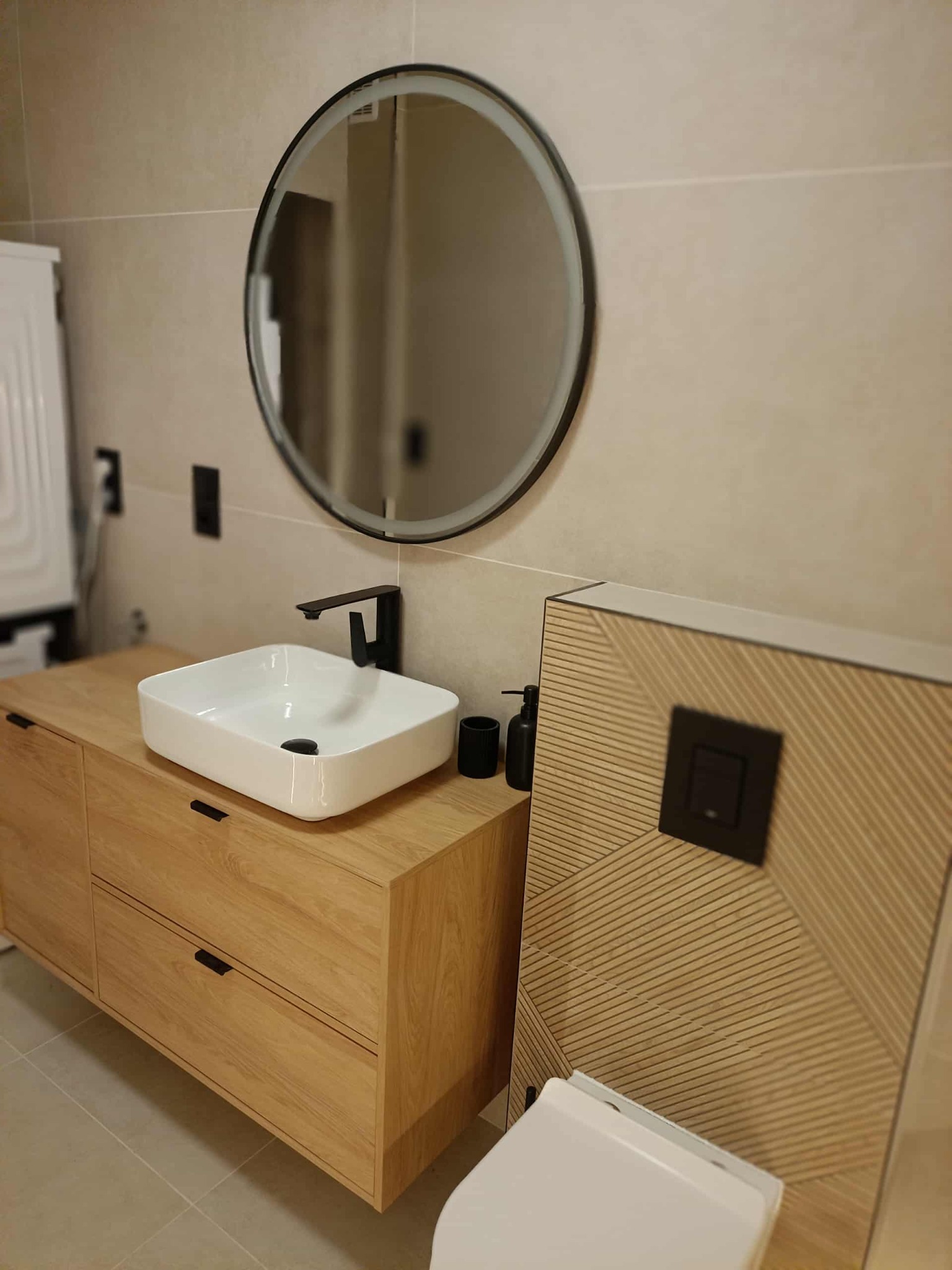 Nowoczesna łazienka w beżach: szafka z umywalką nablatową, okrągłe lustro z czarną ramą i podwieszana toaleta z drewnianym panelem dekoracyjnym.