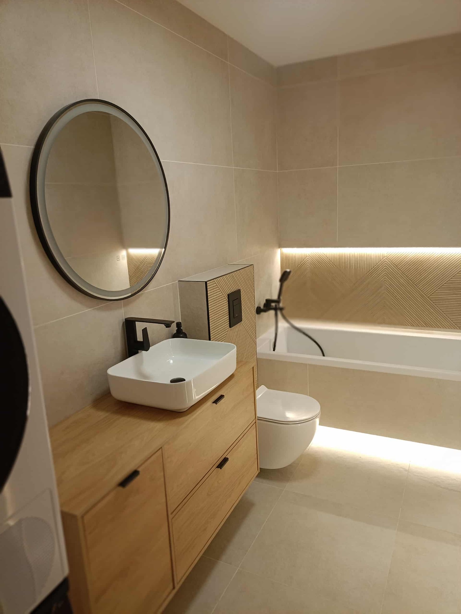 Nowoczesna łazienka w beżowej tonacji z drewnianą szafką pod umywalką, okrągłym lustrem z podświetleniem i podświetlaną wnęką nad wanną.