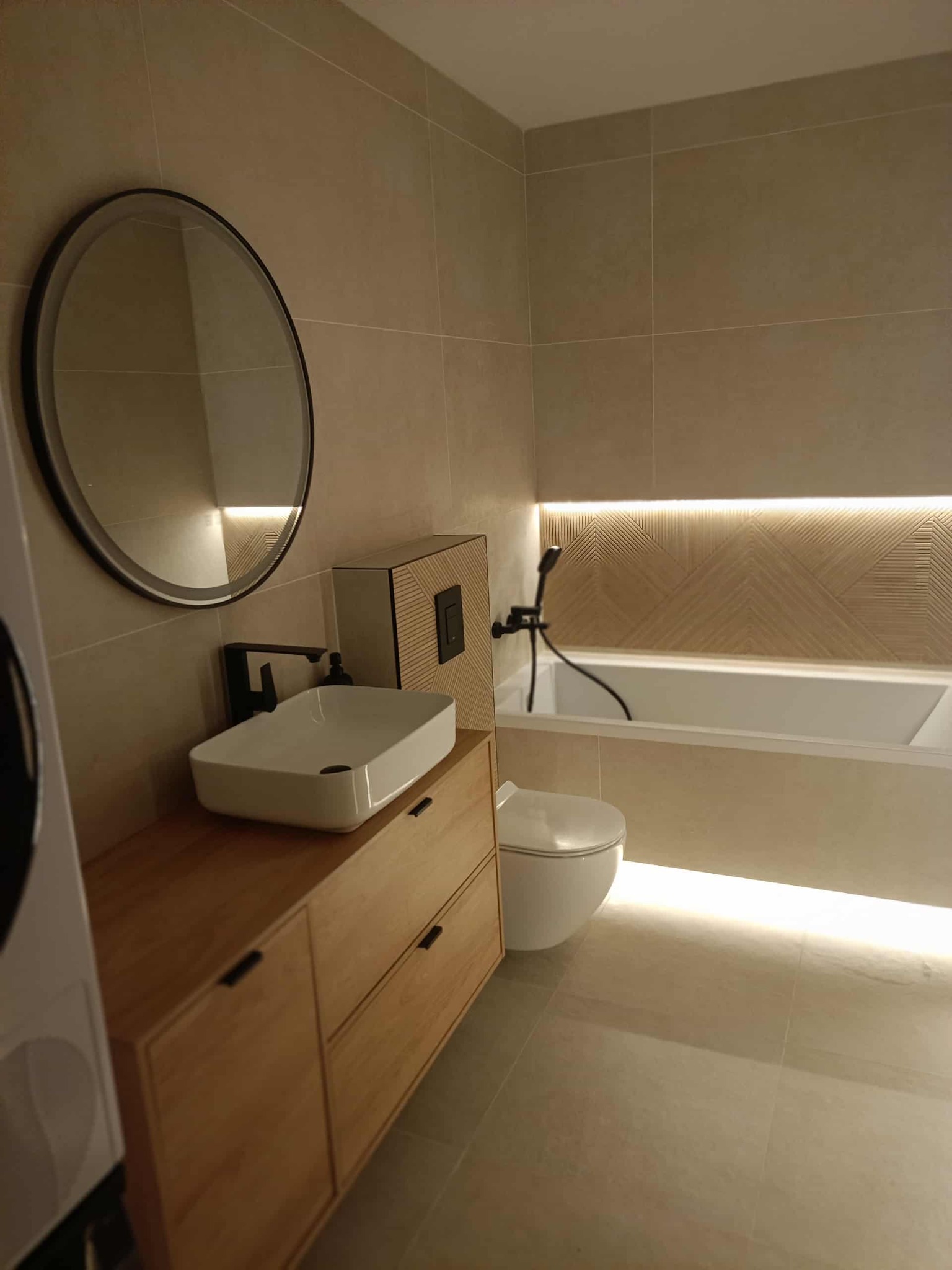 Nowoczesna łazienka w beżowej tonacji z drewnianą szafką pod umywalką, okrągłym lustrem i podświetlaną wanną. Minimalistyczny design i ciepłe oświetlenie.