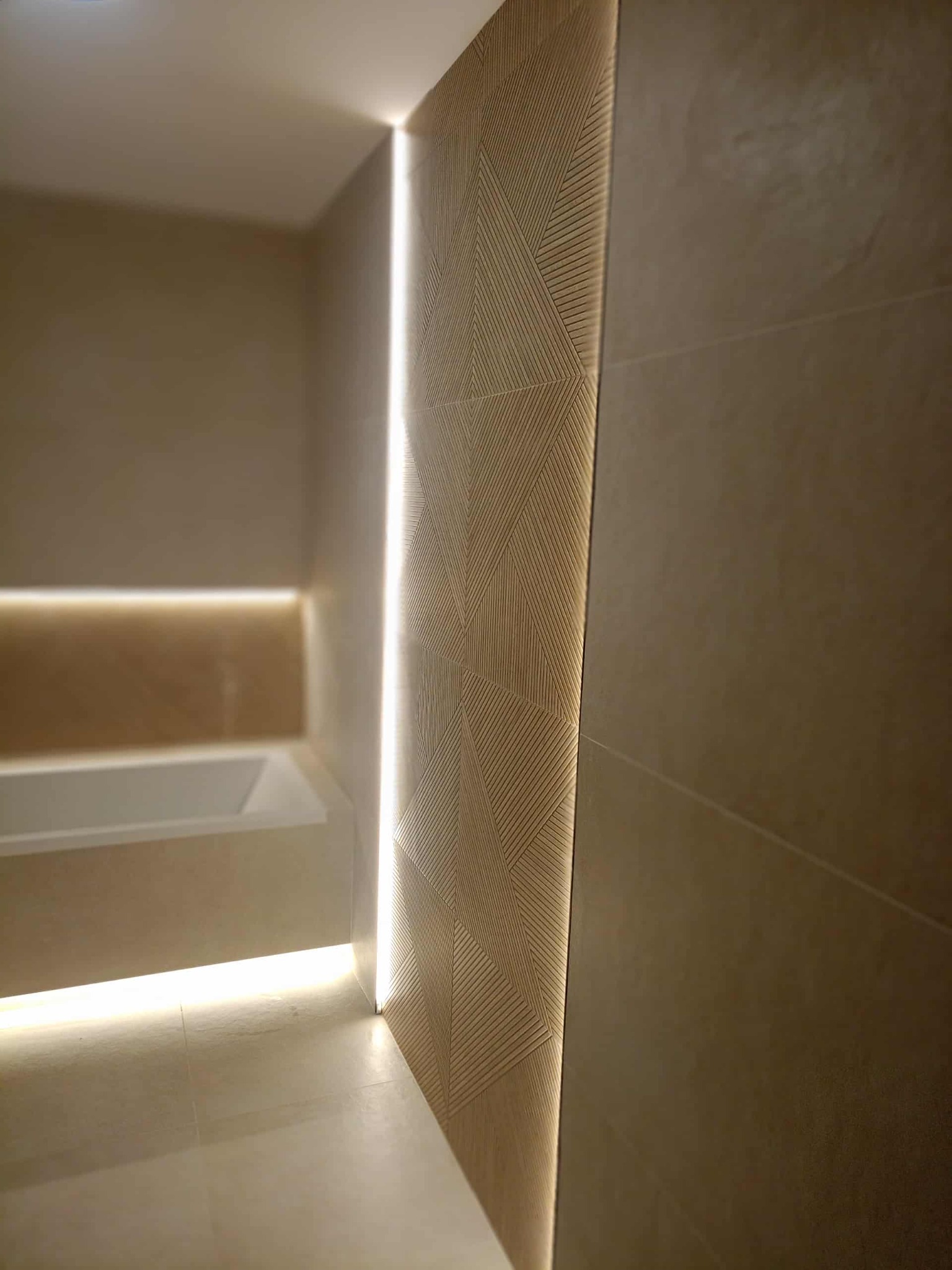 Nowoczesna łazienka z podświetlaną ścianą z geometrycznym wzorem, beżowe płytki, subtelne oświetlenie LED, wanna w tle. Minimalistyczny design.