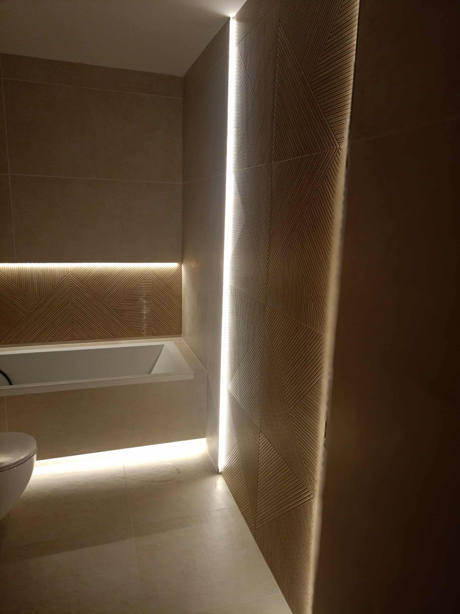 Nowoczesna łazienka z beżowymi płytkami, podświetleniem LED i geometrycznym wzorem na ścianie. Minimalistyczny design z wanną i toaletą w tle.