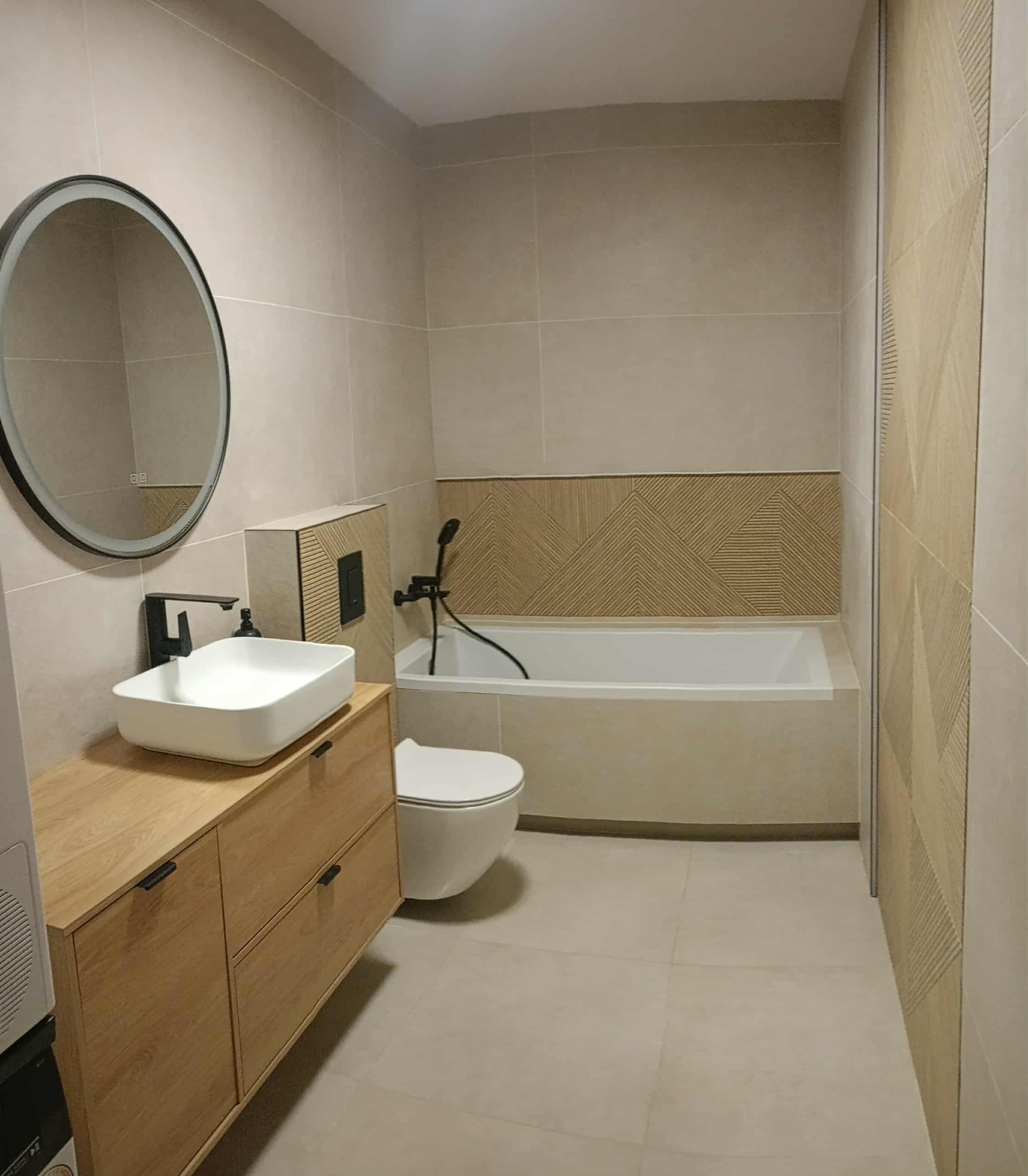 Jasna łazienka z beżowymi płytkami, drewnianą szafką pod umywalką i okrągłym lustrem z podświetleniem. Nad wanną dekoracyjne panele ścienne z motywem jodełki.