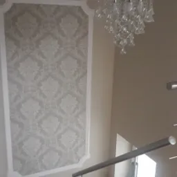 Klatka schodowa z tapetą w ozdobnej ramie, kryształowy żyrandol i metalowa balustrada.