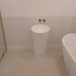 Nowoczesna łazienka z wolnostojącą umywalką w kształcie kolumny, złotą armaturą i fragmentem wanny na tle białych paneli ściennych i beżowych płytek podłogowych.