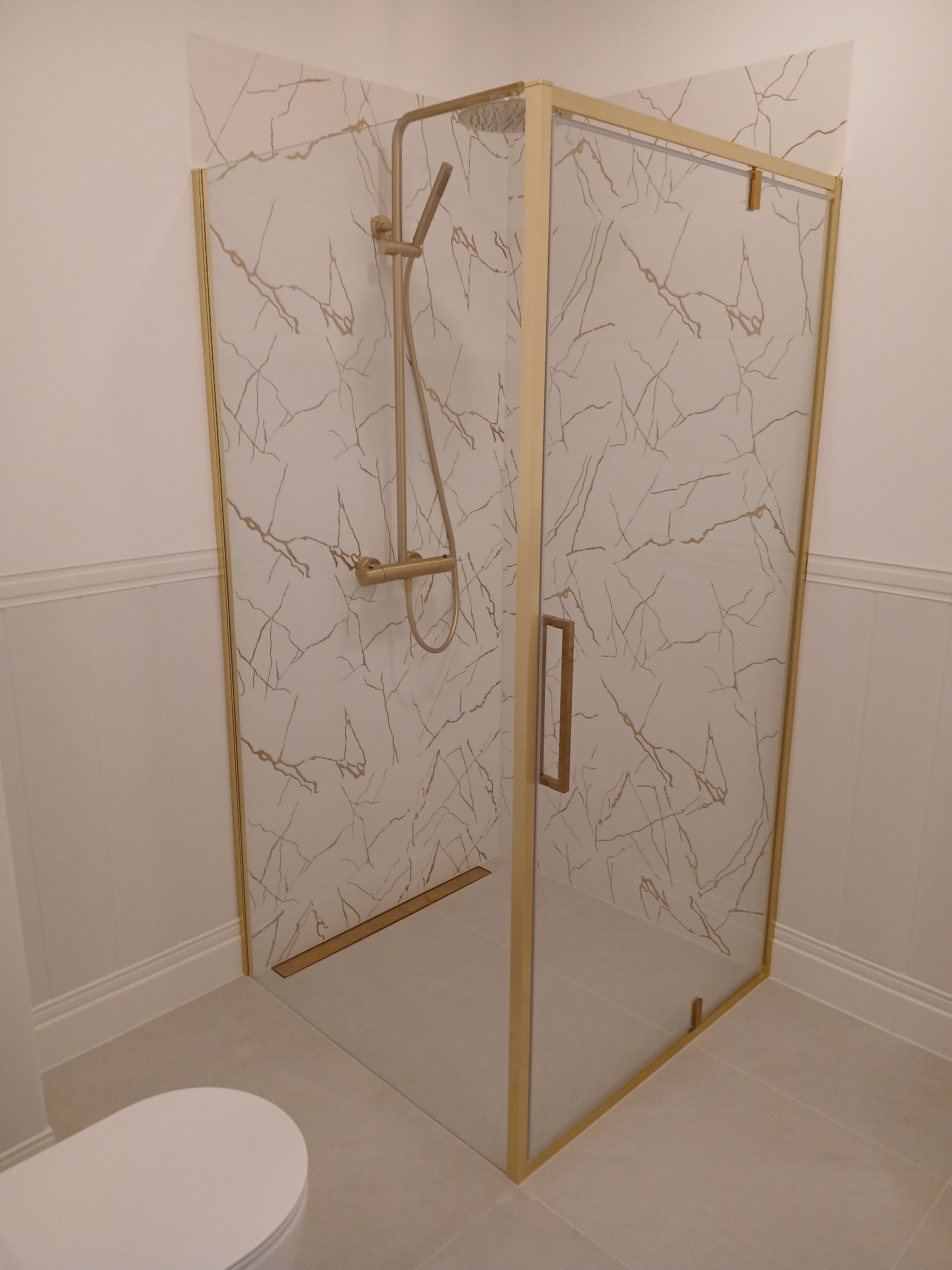 Kabina prysznicowa z elementami w kolorze złotym, płytki imitujące marmur z żyłami w kolorze złotym, widoczna toaleta w tle.