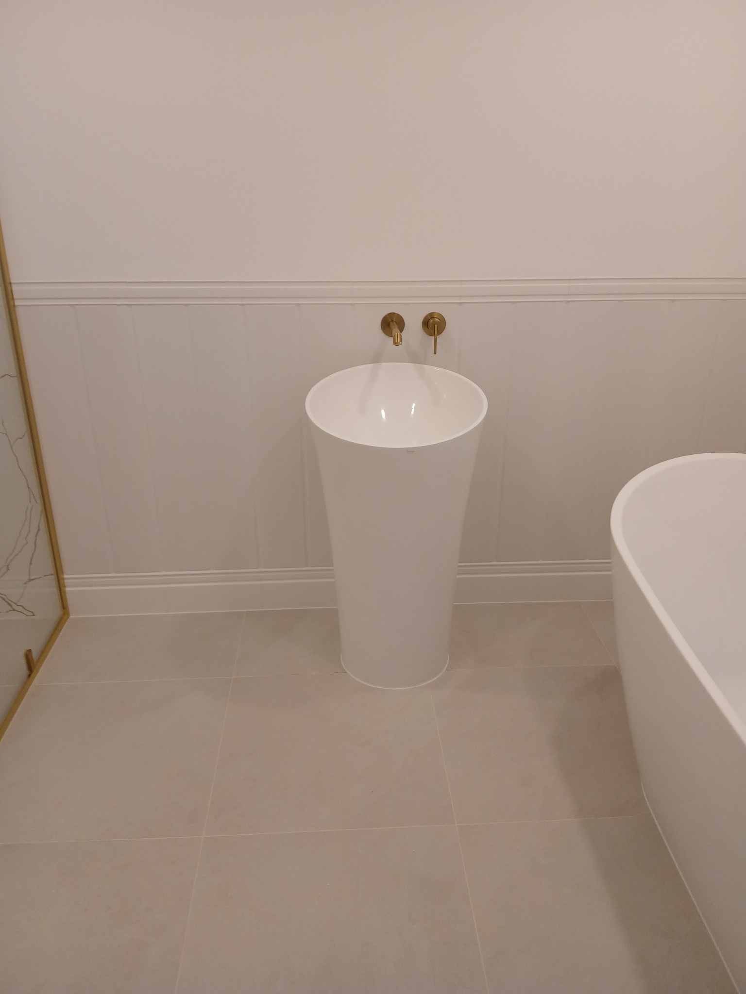 Nowoczesna łazienka z wolnostojącą umywalką w kształcie kolumny, złotą armaturą i fragmentem wanny na tle białych paneli ściennych i beżowych płytek podłogowych.