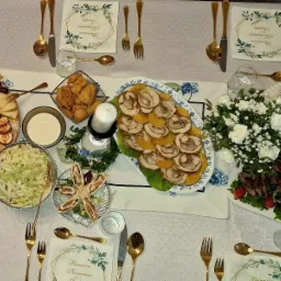 Uroczysty stół komunijny z różnorodnym cateringiem: deska serów, sałatki, roladki z kurczaka na brzoskwiniach, nuggetsy, dekoracje kwiatowe, złote sztućce i serwetki z motywem komunijnym.