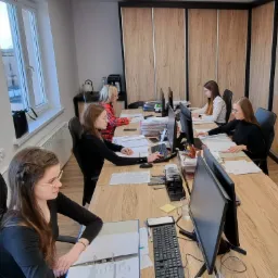 Zespół pracowników biurowych przy biurkach z komputerami i dokumentami, przygotowujących się do rocznych rozliczeń, w jasnym pomieszczeniu z dużym oknem i drewnianymi szafami.