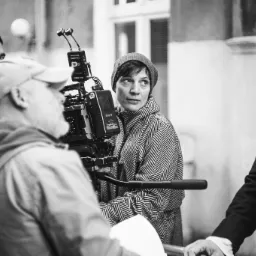 Czarno-białe zdjęcie z planu filmowego: kobieta w czapce i wzorzystej kurtce stoi obok kamery filmowej z mikrofonami, w tle mężczyźni i fragment budynku.