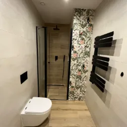 Nowoczesna łazienka z kabiną prysznicową typu walk-in, wiszącą toaletą, grzejnikiem drabinkowym w kolorze czarnym i ścianą wykończoną płytkami z motywem tropikalnym.