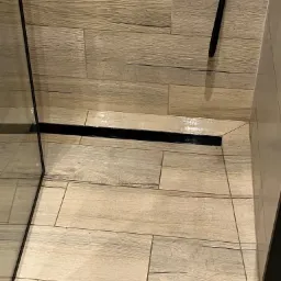 Ułożona pod kątem podłoga z płytek imitujących drewno w kabinie prysznicowej z czarnym odpływem liniowym.