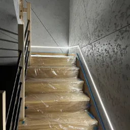 Drewniane schody zabezpieczone folią malarską podczas prac wykończeniowych, z podświetleniem LED wzdłuż ściany o fakturze betonu architektonicznego.