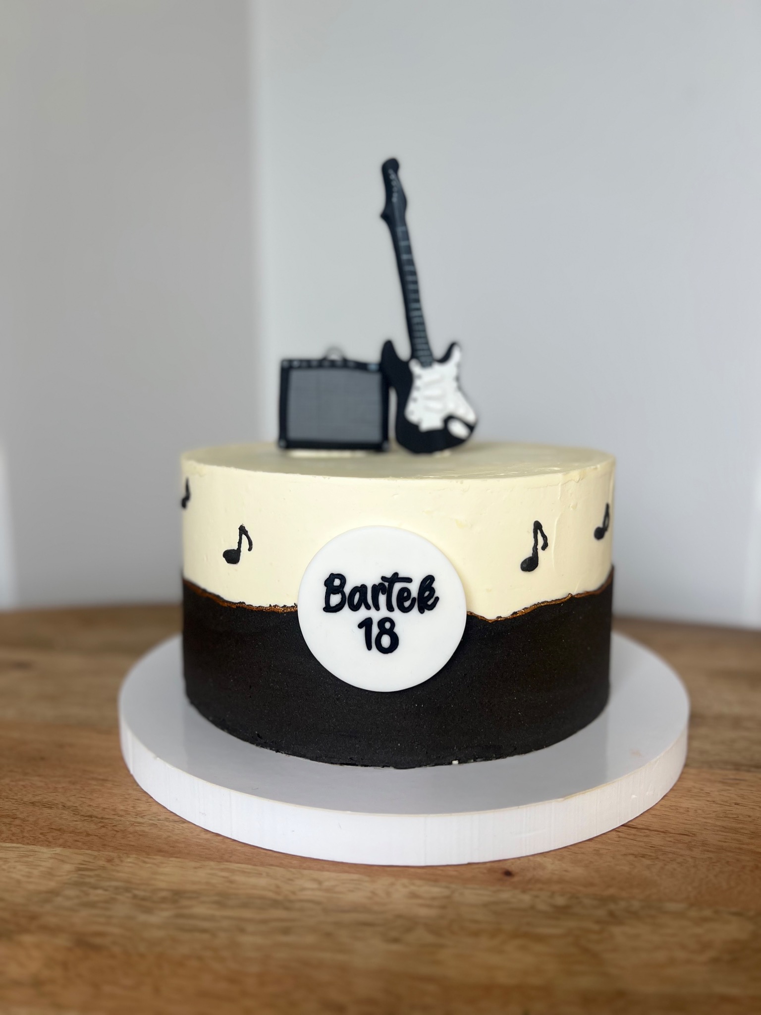 Tort dwukolorowy z dekoracjami muzycznymi: gitara, wzmacniacz i nuty, z napisem 'Bartek 18' na białym tle, prezentowany na drewnianym blacie.