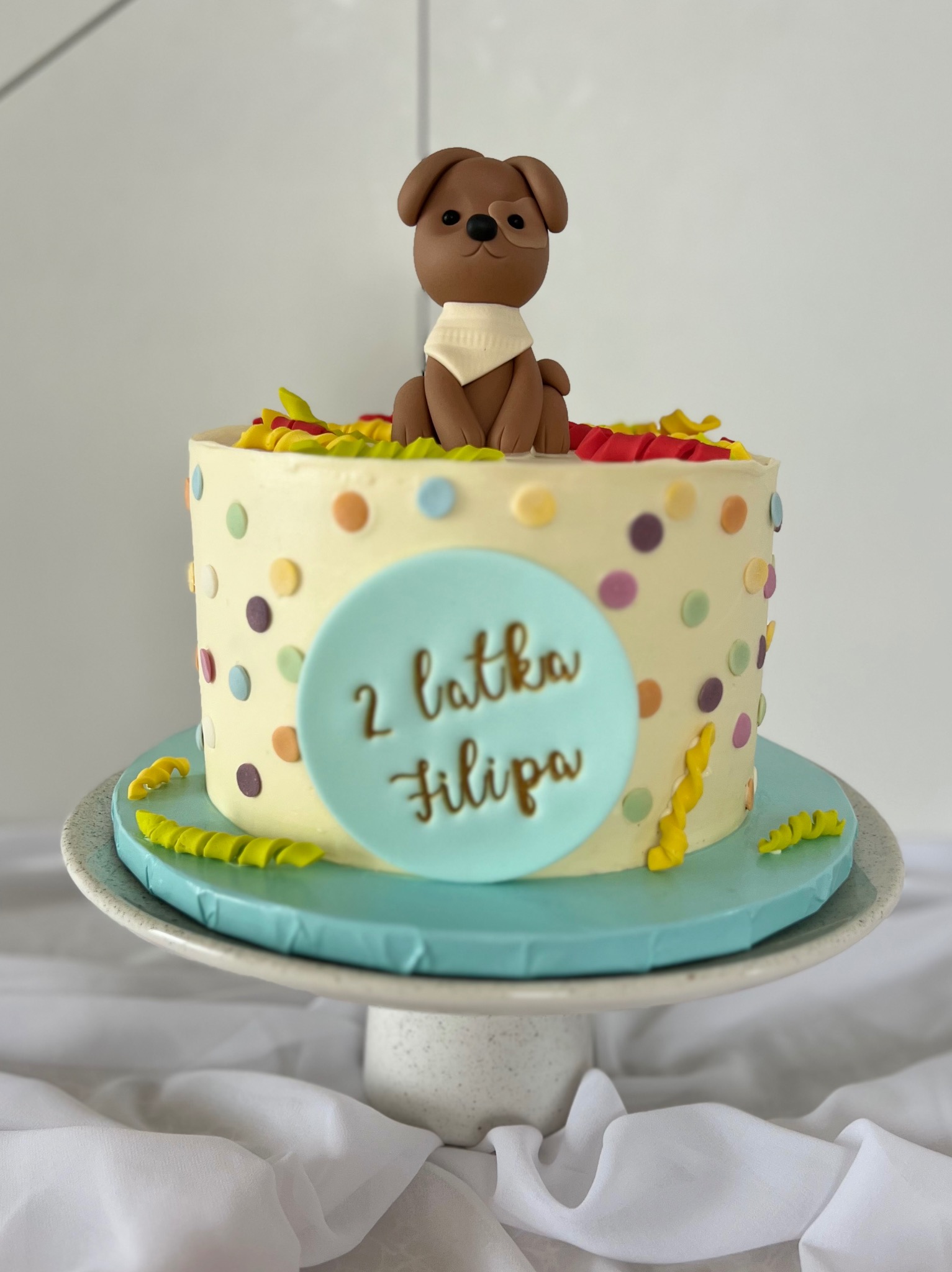Tort urodzinowy z figurką psa z masy cukrowej i napisem '2 latka Filipa', wykonany w Wejherowie, na paterze z motywem confetti.
