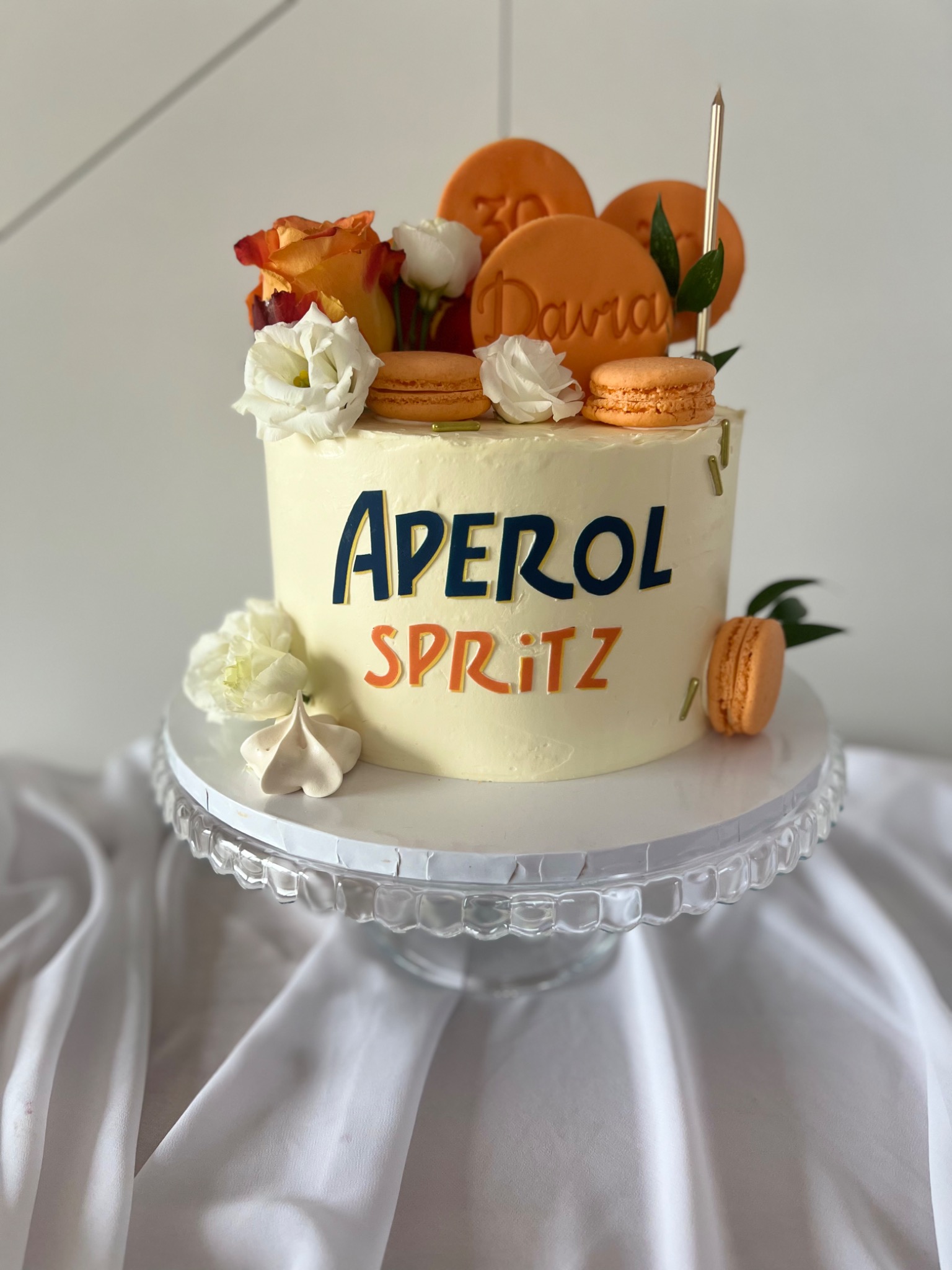 Tort z kremem, dekorowany kwiatami, makaronikami, okrągłymi ciasteczkami z napisem 'Daria 30' oraz napisem 'Aperol Spritz', umieszczony na szklanej paterze na białym obrusie.
