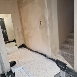 Wnętrze w trakcie prac wykończeniowych: ściany po nałożeniu tynku, widoczne przewody elektryczne, podłoga zabezpieczona folią i przygotowana do dalszych prac, w tle schody w stanie surowym.