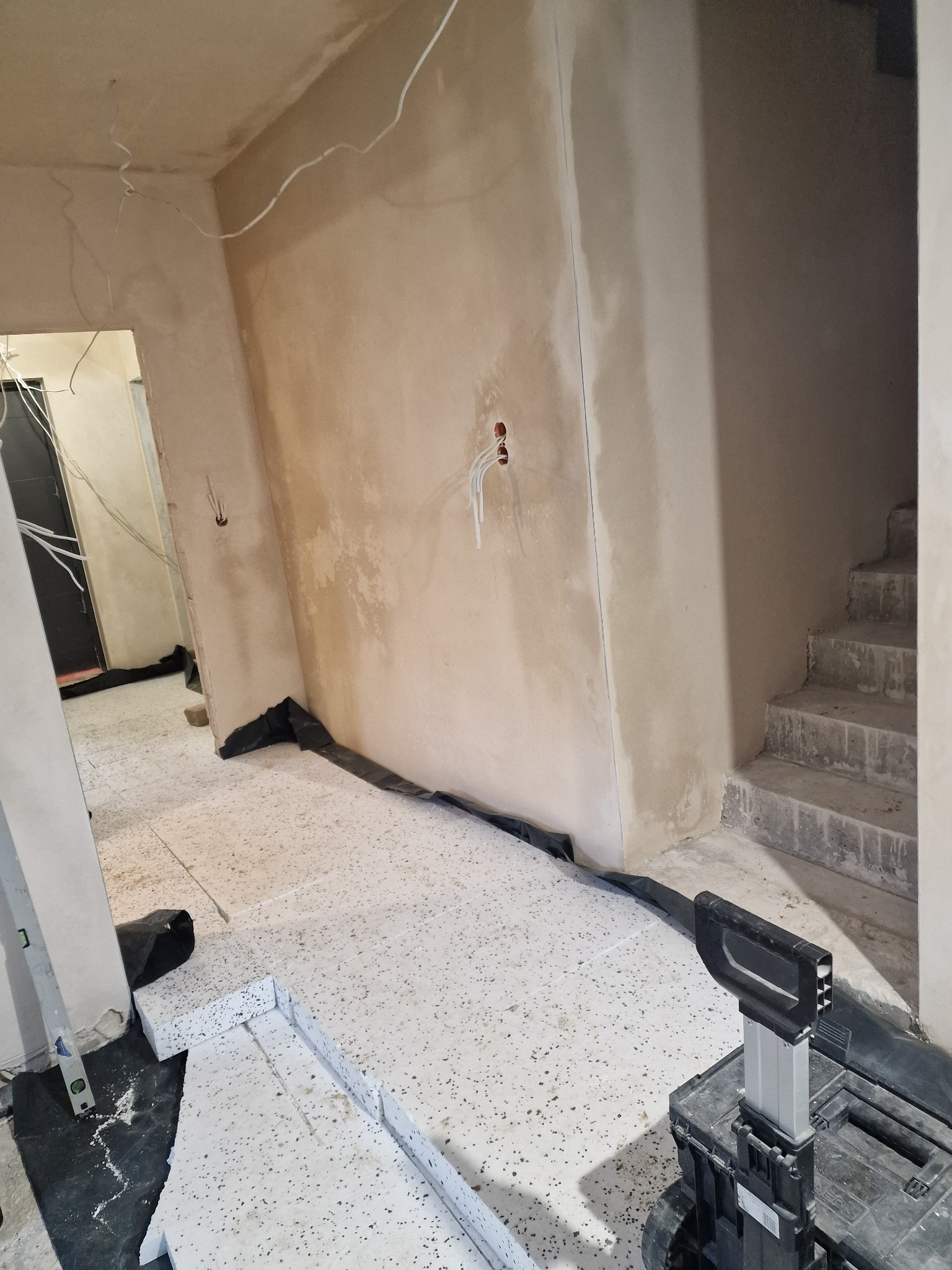 Wnętrze w trakcie prac wykończeniowych: ściany po nałożeniu tynku, widoczne przewody elektryczne, podłoga zabezpieczona folią i przygotowana do dalszych prac, w tle schody w stanie surowym.