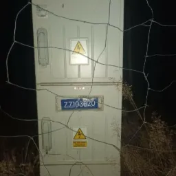 Szara skrzynka elektryczna na zewnątrz, widoczna przez siatkę ogrodzeniową, z ostrzeżeniami o wysokim napięciu i numerem identyfikacyjnym Z7103820.