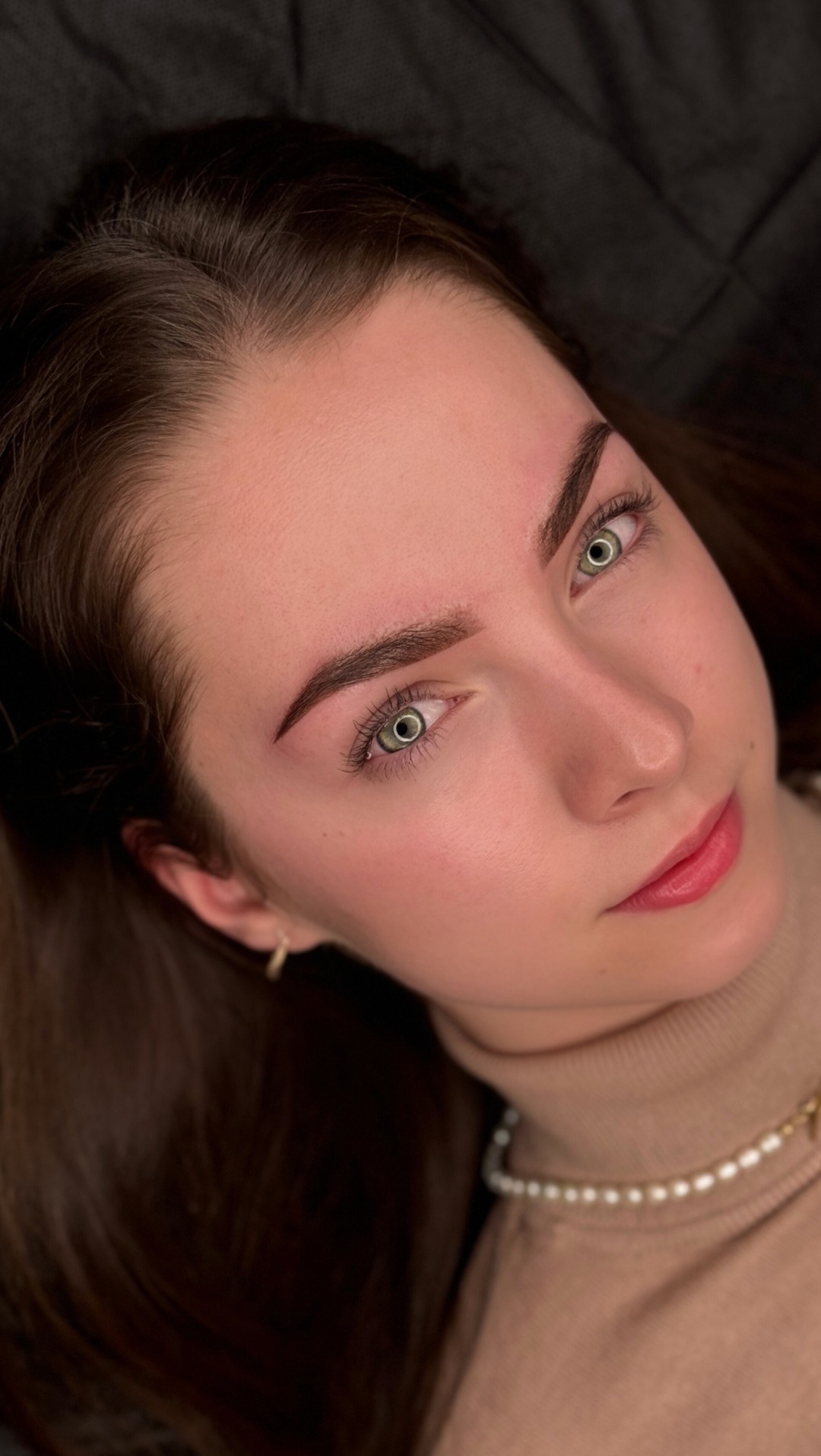 Portret młodej kobiety z zielonymi oczami i świeżo wystylizowanymi brwiami, widoczne delikatne włoski i precyzyjny kształt, ujęcie z góry w naturalnym świetle, widoczny złoty kolczyk i perłowy...