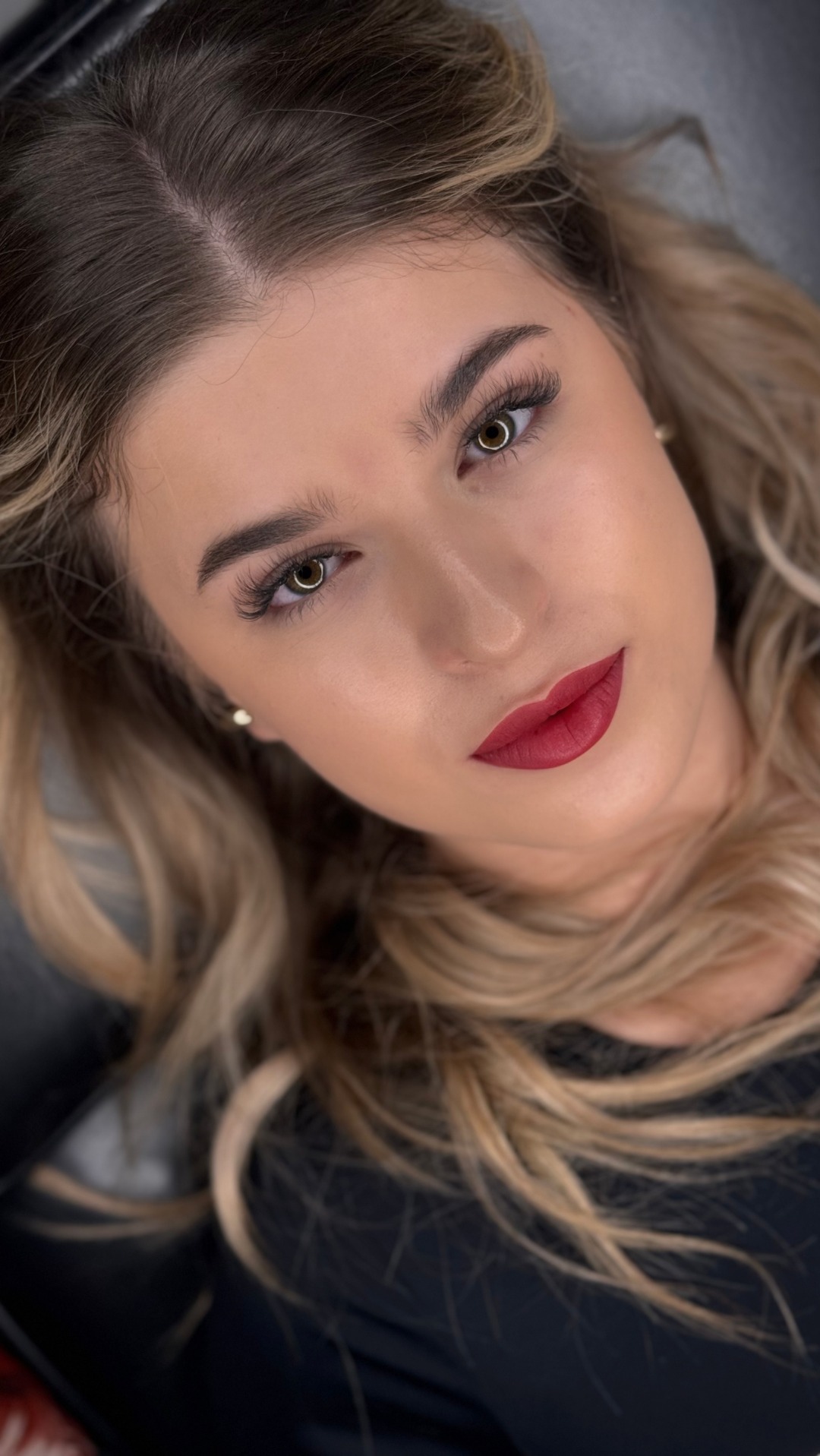 Portret kobiety z profesjonalnym, pełnym makijażem: intensywna czerwona szminka, podkreślone brwi i rzęsy, delikatny makijaż oczu, blond włosy ułożone w fale.