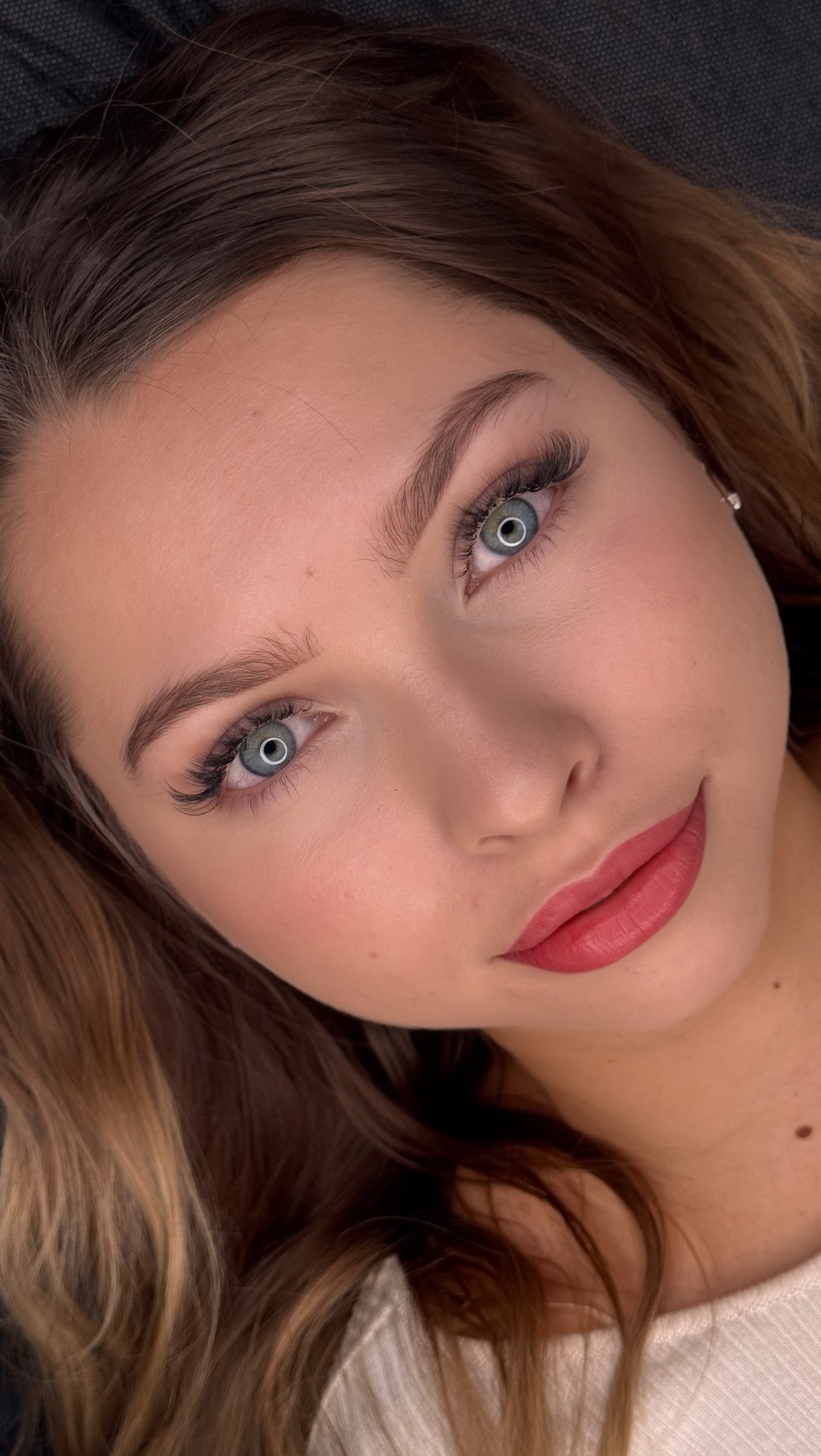 Portret kobiety z intensywnym spojrzeniem, podkreślonymi rzęsami i pomalowanymi ustami w odcieniu koralowej czerwieni. Widoczny delikatny rumieniec na policzkach i subtelny blask na powiekach.