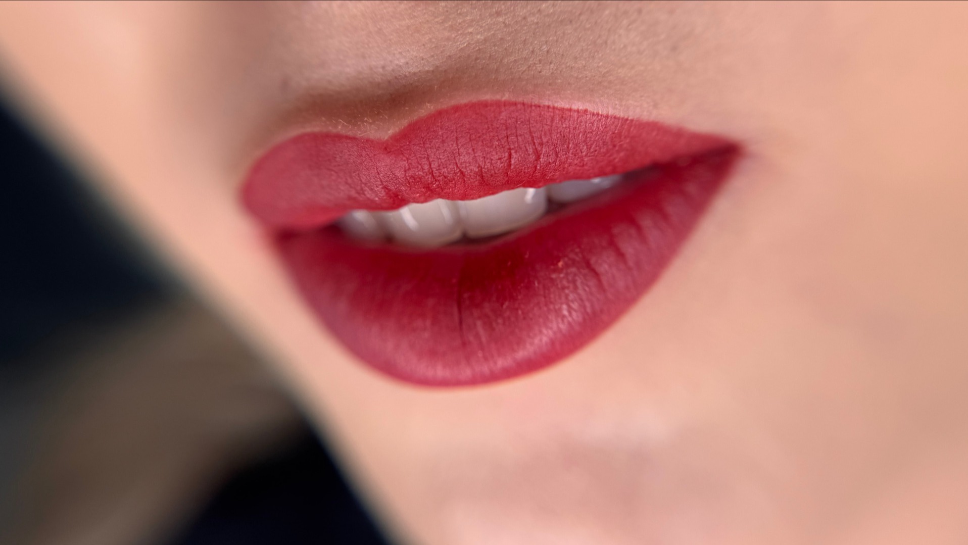 Zbliżenie na usta z intensywnym czerwonym makijażem, delikatny uśmiech eksponuje białe zęby, gładka skóra w tle.
