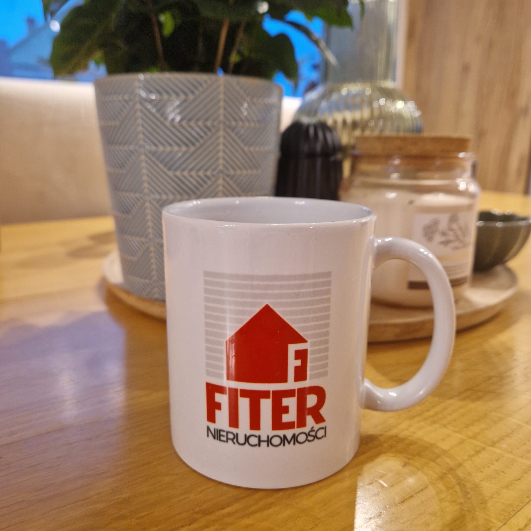 Kubek z logo 'FITER Nieruchomości' na drewnianym stole, obok doniczki i świeczki, w ciepłym oświetleniu. Dominuje biel i brąz, tworząc przytulną atmosferę.