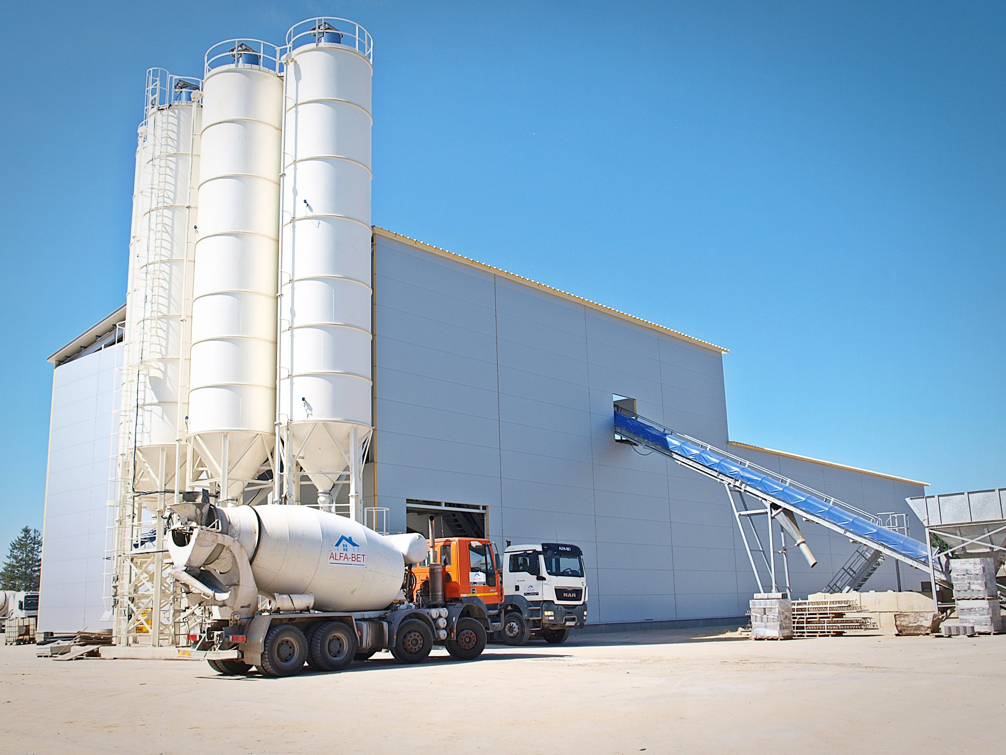 Widok zewnętrzny betoniarni z silosami, taśmociągiem i betonomieszarką firmy Alfa-Bet, stojącą obok pomarańczowej ciężarówki MAN.