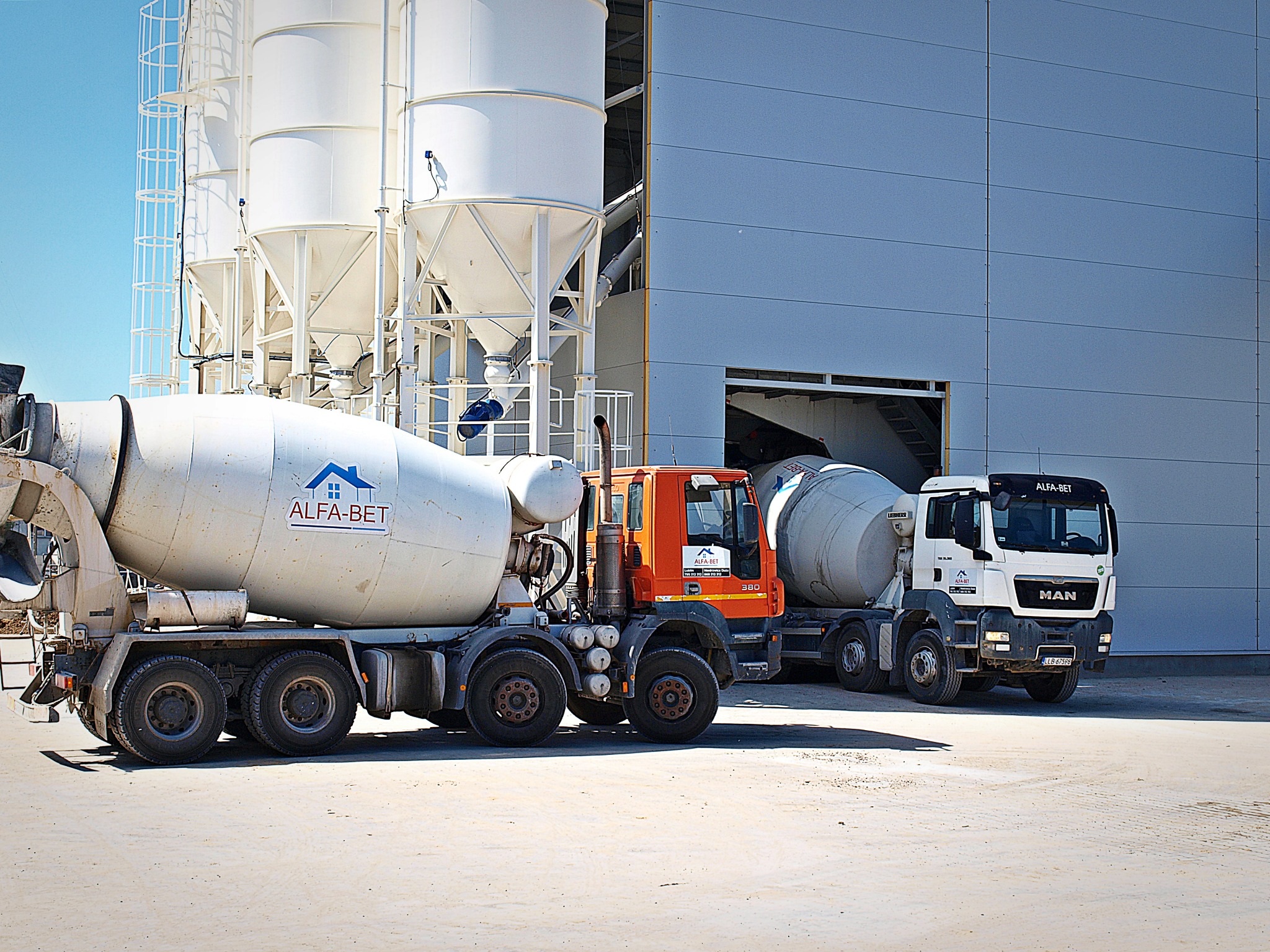 Dwa betonomieszarki Alfa-Bet, jeden pomarańczowy i jeden biały, stojące przed silosami i budynkiem przemysłowym w słoneczny dzień.