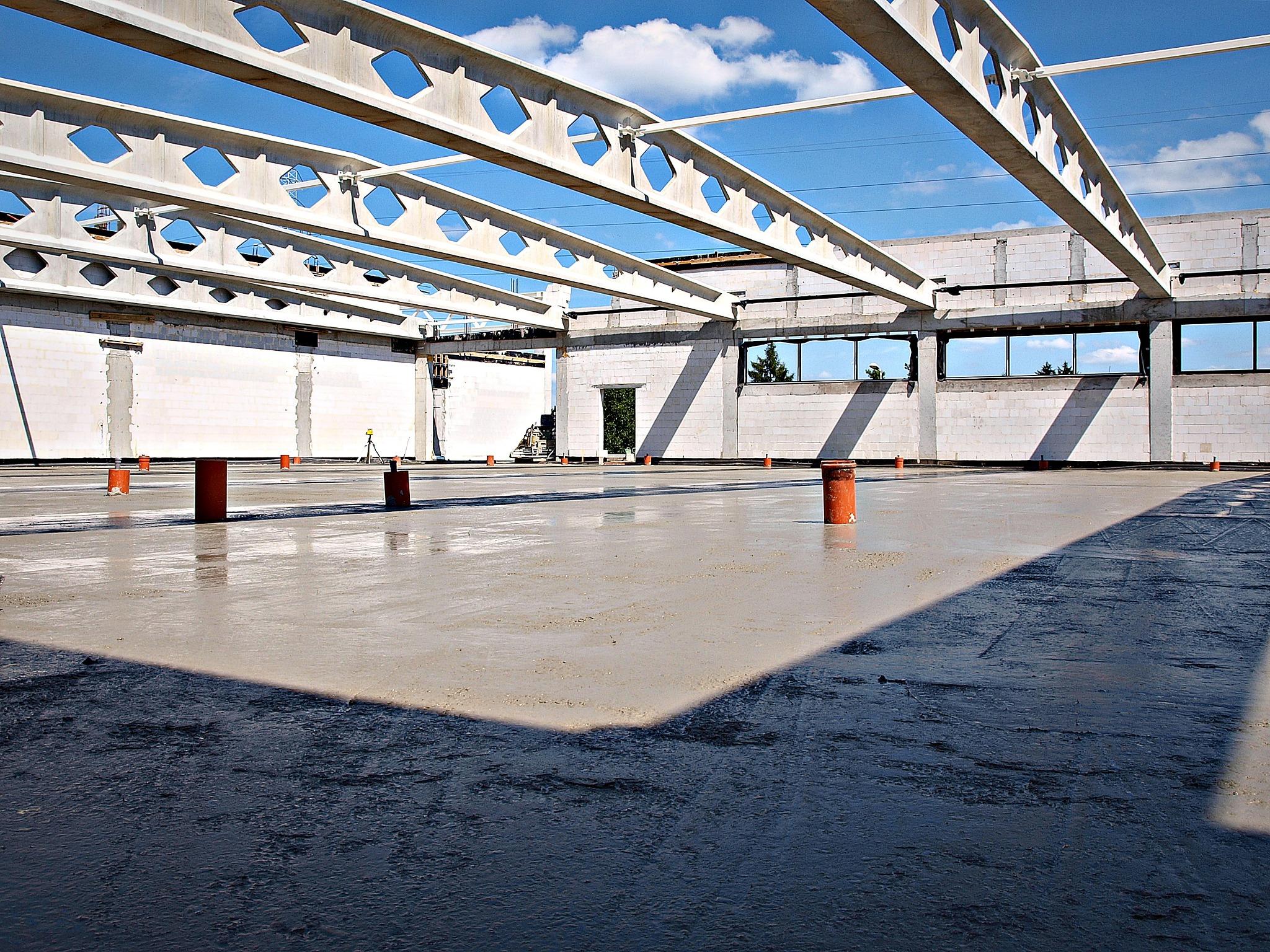 Świeżo wylana, mokra posadzka w hali przemysłowej z widocznymi stalowymi belkami stropowymi o ażurowej konstrukcji i rzędami okien w tle, oświetlona naturalnym światłem słonecznym.