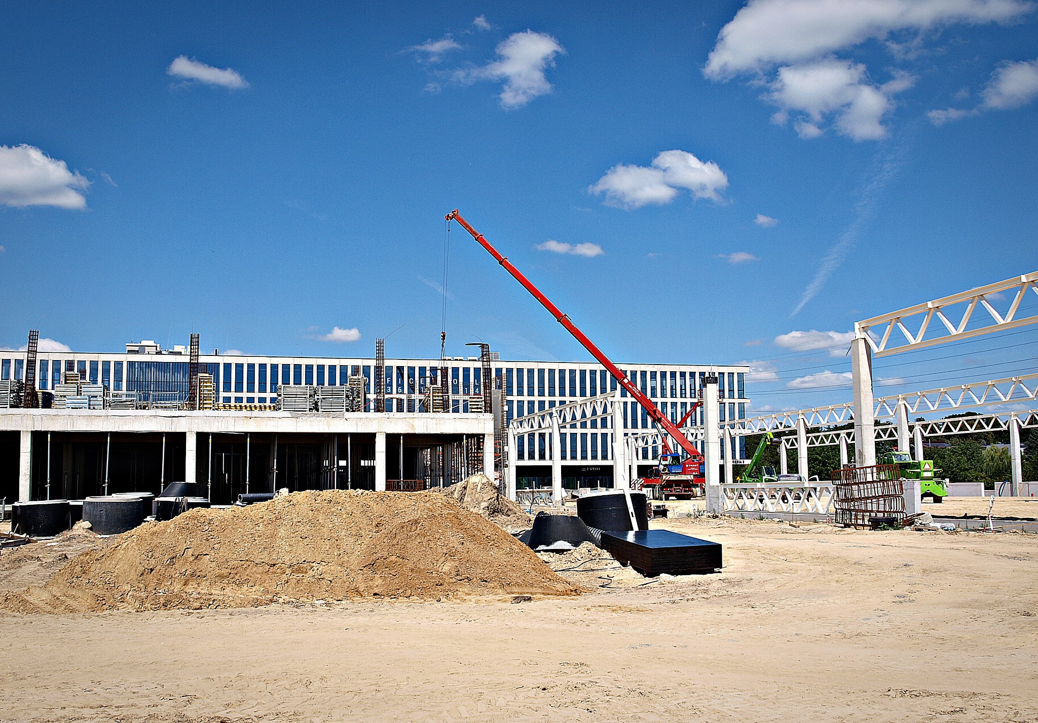 Plac budowy z czerwoną żurawiem, stertą piasku, betonowymi prefabrykatami i fragmentem budynku w stanie surowym.