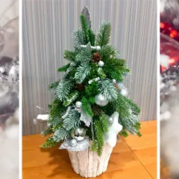 Dekoracyjna choinka z zielonych gałązek, srebrnymi ozdobami, szyszkami i białą gwiazdą w teksturowanej, jasnej doniczce na drewnianym stole, z rozmytym tłem z bombkami.