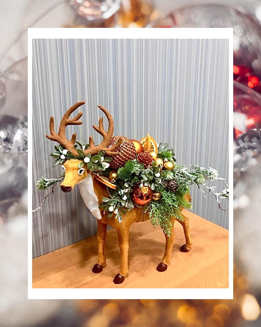 Dekoracja świąteczna: filcowy renifer z porożem ozdobionym igliwiem, bombkami i suszonymi pomarańczami na drewnianym blacie.