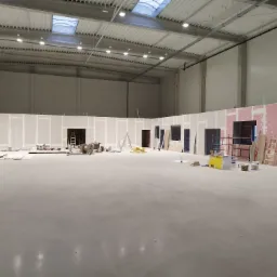 Wnętrze hali produkcyjnej w trakcie adaptacji: widoczne ścianki działowe z płyt gipsowo-kartonowych, otwory drzwiowe, drabiny, materiały budowlane i narzędzia, betonowa posadzka, oświetlenie sufitowe.