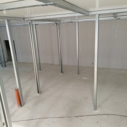 Artur Sarnecki - Montaż stelażu sufitu podwieszanego z profili metalowych, widoczne białe panele sufitowe i pomarańczowa rura w rogu pomieszczenia.